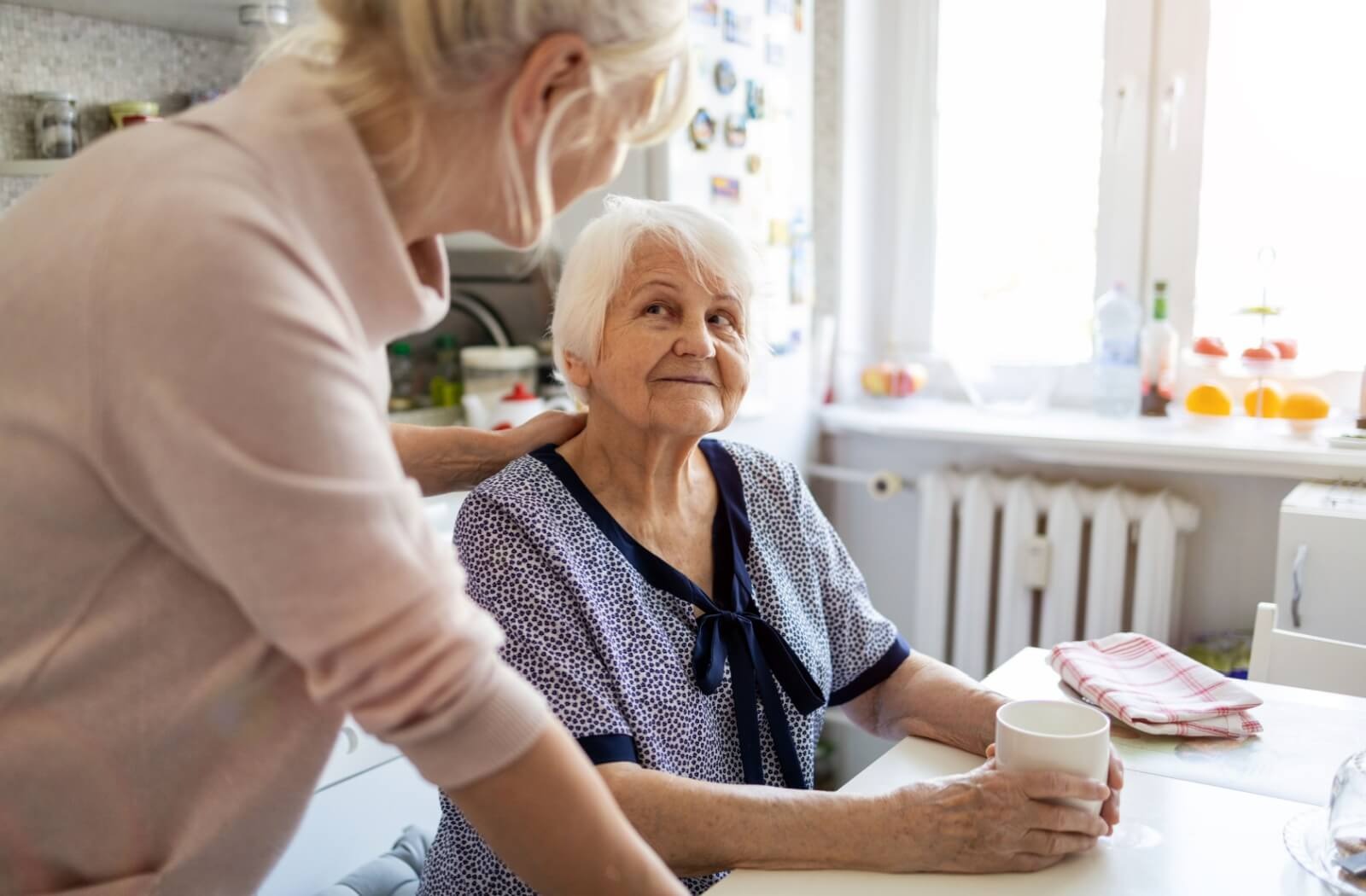 An adult places their hand on their senior parent's shoulder, who is sitting at a table with a coffee cup.