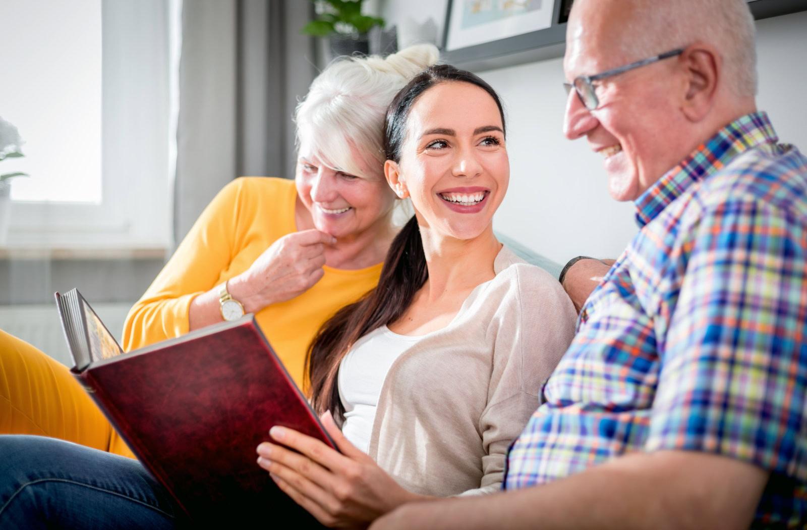 two seniors and a young adult laugh on a couch while reading a photo album
